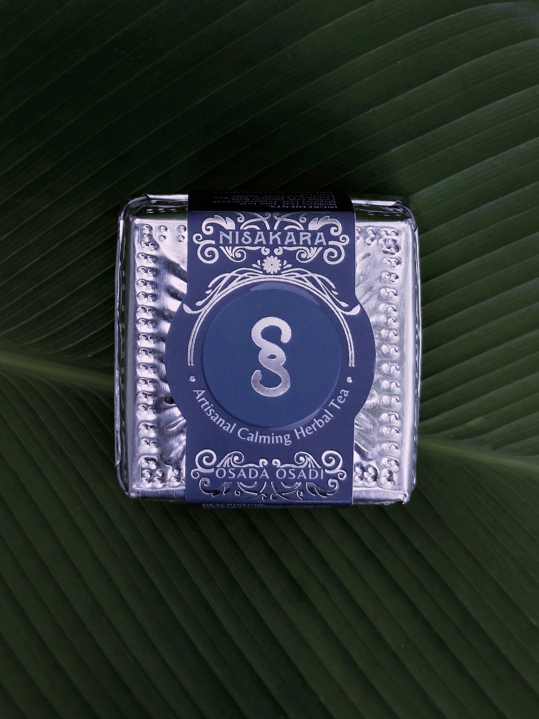 Nisakara artisanal calming herbal tea is packaged in a silver tea tin from OSADA OSADI, sitting on a leaf.